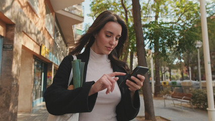 Beautiful young woman walking down the street with grocery bag and using mobile phone