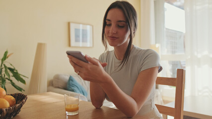 Smiling young woman scrolling through photo on her mobile phone while sitting in the kitchen