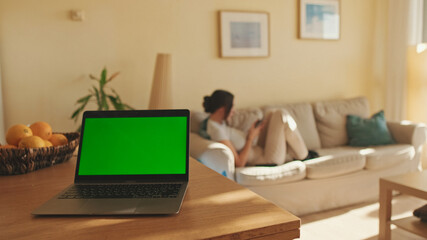 Foreground on the table is laptop with green screen chroma key, in the background girl is sitting,...