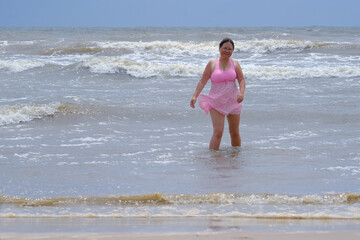 mature woman 50 years old with long hair in pink swimsuit comes out water to seashore, concept swim in waves, dangers of rip currents, safety rules in sea, travel, vacation in tropical, sun protection
