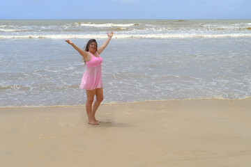 woman 50 years old with long hair in pink swimsuit stands barefoot on sandy seashore, enjoys sea...