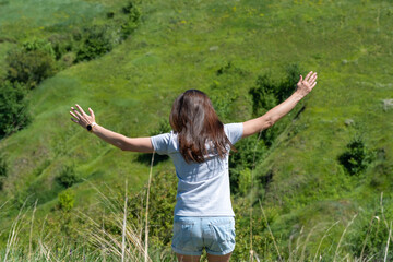 Happy woman greeting sun in ravine covered with greenery. Young female raise hands to the sunshine. She looking on valley and showing  freedom gesture. Positive joyful of emotion surya namaskar.