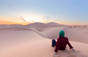 A young male traveler enjoys a sunrise or sunset landscape view of the desert sand dunes of Erg...