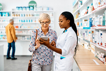 Plakat Senior woman and black female pharmacist using digital tablet in drugstore.