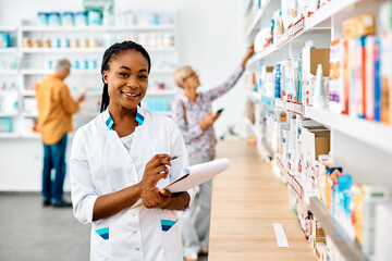 Happy black female pharmacist taking inventory in pharmacy and looking at camera.