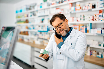 Happy pharmacist holding medicine while talking on mobile phone in pharmacy.
