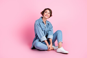 Full length photo of shiny thoughtful woman wear jeans shirt sitting floor looking empty space isolated pink color background