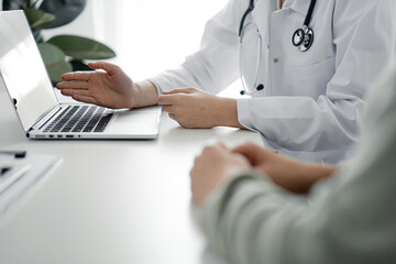 Doctor and patient sitting at the desk in clinic office. The focus is on female physician's hands pointing to laptop computer monitor, close up. Medicine concept