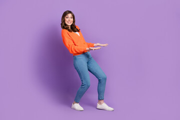 Full length photo of impressed funny woman dressed orange cardigan carrying arms empty space isolated purple color background