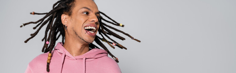 positive multiracial man with dreadlocks shaking head and smiling isolated on grey, banner