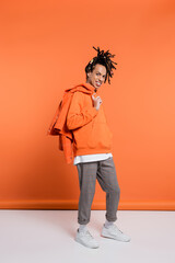 Fototapeta premium full length of positive multiracial man in hoodie holding denim jacket on coral background