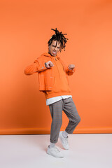 Fototapeta premium full length of excited and multiracial man with dreadlocks standing with clenched fists on coral background