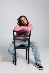 full length of pensive multiracial man with dreadlocks sitting on chair on grey