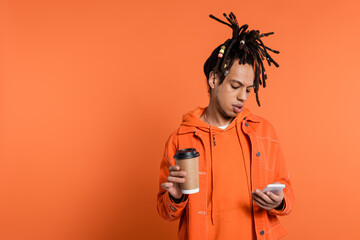 pierced and multiracial man with dreadlocks holding paper cup and using smartphone on coral background
