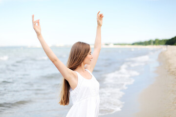 Fototapeta na wymiar Happy smiling woman in free happiness bliss on ocean beach standing with raising hands. Portrait of a multicultural female model in white summer dress enjoying nature 