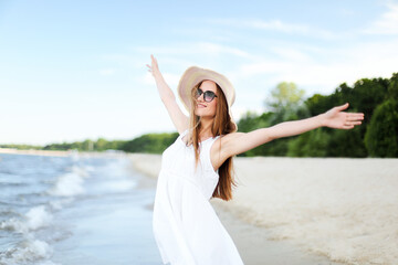 Fototapeta na wymiar Happy smiling woman in free happiness bliss on ocean beach standing with a hat, sunglasses, and raising hands. Portrait of a multicultural female model in white summer dress enjoying 