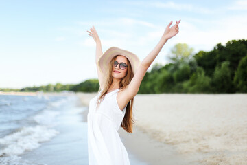 Happy smiling woman in free happiness bliss on ocean beach standing with a hat, sunglasses, and raising hands. Portrait of a multicultural female model in white summer dress enjoying 