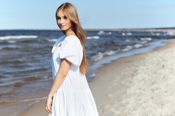 Happy, beautiful woman on the ocean beach standing in a white summer dress. Portrait