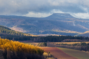 A view of the Szczeliniec Mountain Peak, towering over the Kłodzko Valley. Autumn trees illuminated by the sun. - obrazy, fototapety, plakaty