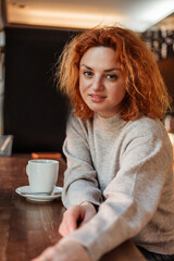 Red-haired curly young bright woman with green eyes drinks hot coffee in cozy cafe. Beautiful model in stylish clothes. Europe.