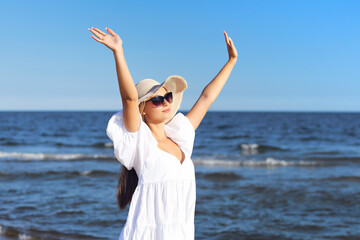 Happy blonde woman is on the ocean beach in a white dress and sunglasses, raising hands