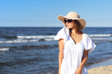 Happy smiling blonde woman is posing on the ocean beach with sunglasses and a hat