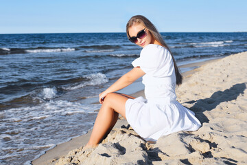 Fototapeta na wymiar Happy, beautiful blonde woman is sitting on the ocean beach in a white summer dress and sunglasses