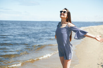 Happy smiling woman in free bliss on ocean beach standing with open hands. Portrait of a brunette female model in summer dress enjoying nature during travel holidays vacation outdoors