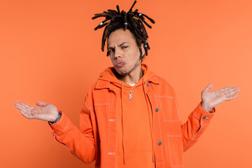 tattooed and multiracial man with dreadlocks showing shrug gesture isolated on coral background