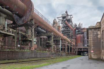 huge pipes of an old industrial plant lead through the picture. It is dominated by rusty brown colors. On the ground a small strip of green.