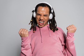 excited multiracial man with dreadlocks listening music in wireless headphones and showing yes gesture isolated on grey