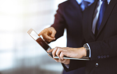 Business people discussing a presentation at meeting, standing in a sunny modern office. Unknown businessman with a colleague search for some information at the laptop, lawyers at negotiation