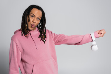 multiracial man with dreadlocks pouting lips while holding wireless headphones on grey