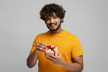 Young smiling confident Indian man holding gift box looking at camera isolated on gray background. Holidays, happy birthday concept  