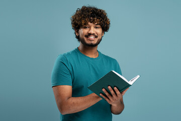 Portrait of handsome smiling Indian man reading book isolated on blue background. Smart happy asian student studying, learning language, exam preparation looking at camera. Education concept  