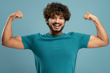 Strong smiling Indian man showing muscles, biceps looking at camera isolated on blue background....