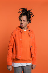 pierced multiracial man with dreadlocks pouting lips while standing in denim jacket on coral background