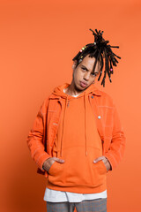 pierced and multiracial man with dreadlocks looking at camera while standing with hands in pockets on coral background