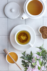 Buckwheat tea in a white cup stands on a saucer, next to a teapot, buckwheat and honey on a white background with flowers.
