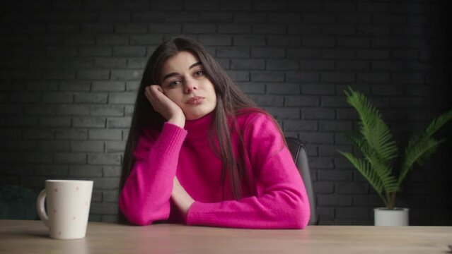 The girl sits at the table with a cup of coffee and thinks how to solve a problem. Bored woman in a pink sweater props her head up with her hand.