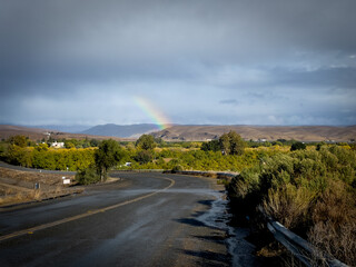 Rainbow touching down on the hills over the orchard beyond the winding road. Wet asphalt road curving to the left toward the rainbow in the sky with the gray storm clouds right after the rain