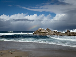 Fototapeta na wymiar Rainbow on horizon with storm clouds in the sky with waves crashing off shore on rock formation at beach in Monterey County, California