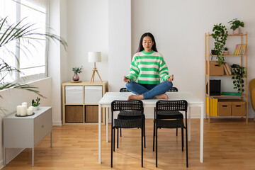 Peaceful young adult asian woman meditating sitting on the table in the living room at home. Chinese trendy female in lotus position practicing meditation exercise indoors