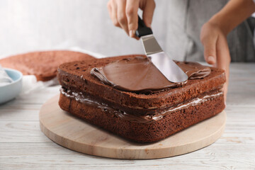 Woman smearing sponge cake with chocolate cream at white wooden table, closeup