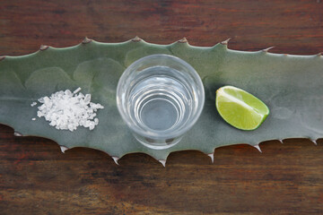 Mexican tequila shot, salt, lime slice and green leaf on wooden table, top view. Drink made of agava
