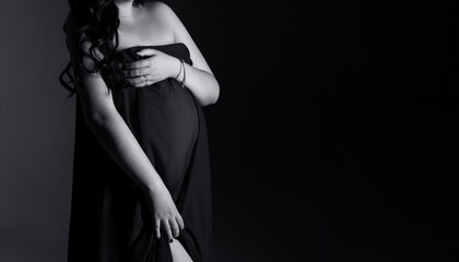 Pregnant woman in a dress standing on a black background with copy space. Pregnancy woman holding her belly.