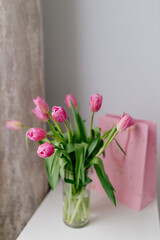 A bouquet of Pink tulips in a glass vase, a pink gift bag stand in a bright room. Background for Valentine's Day. Gift for Mother's Day or Women's Day. Spring background