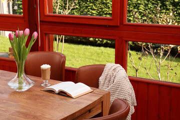 Fototapeta premium Glass of delicious cocoa, pink tulips and book on wooden table at terrace