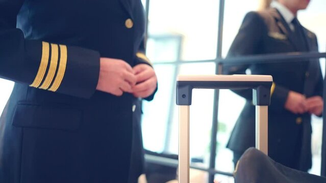 Slow motion of female pilot fastening coat buttons near mirror before departure, pilot in uniform and looking in mirror standing near suitcase in hotel room before flight