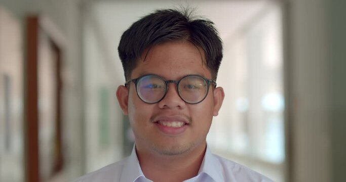 A little smile of an Asian male teenage high school student wearing thick glasses standing on the corridor in the building.